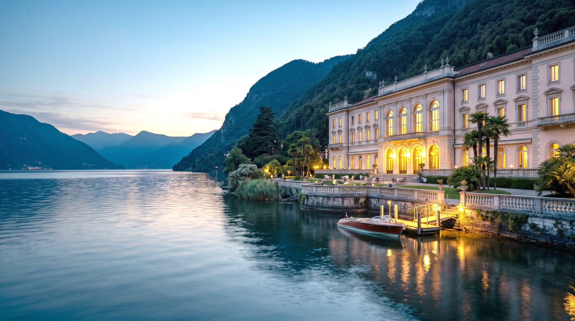Un grande albergo storico sulla riva di un lago del nord Italia al crepuscolo: la lunga facciata chiara con le finestre accese, la terrazza con la balaustra, il pontile con un motoscafo in legno e le montagne che si specchiano nell'acqua.